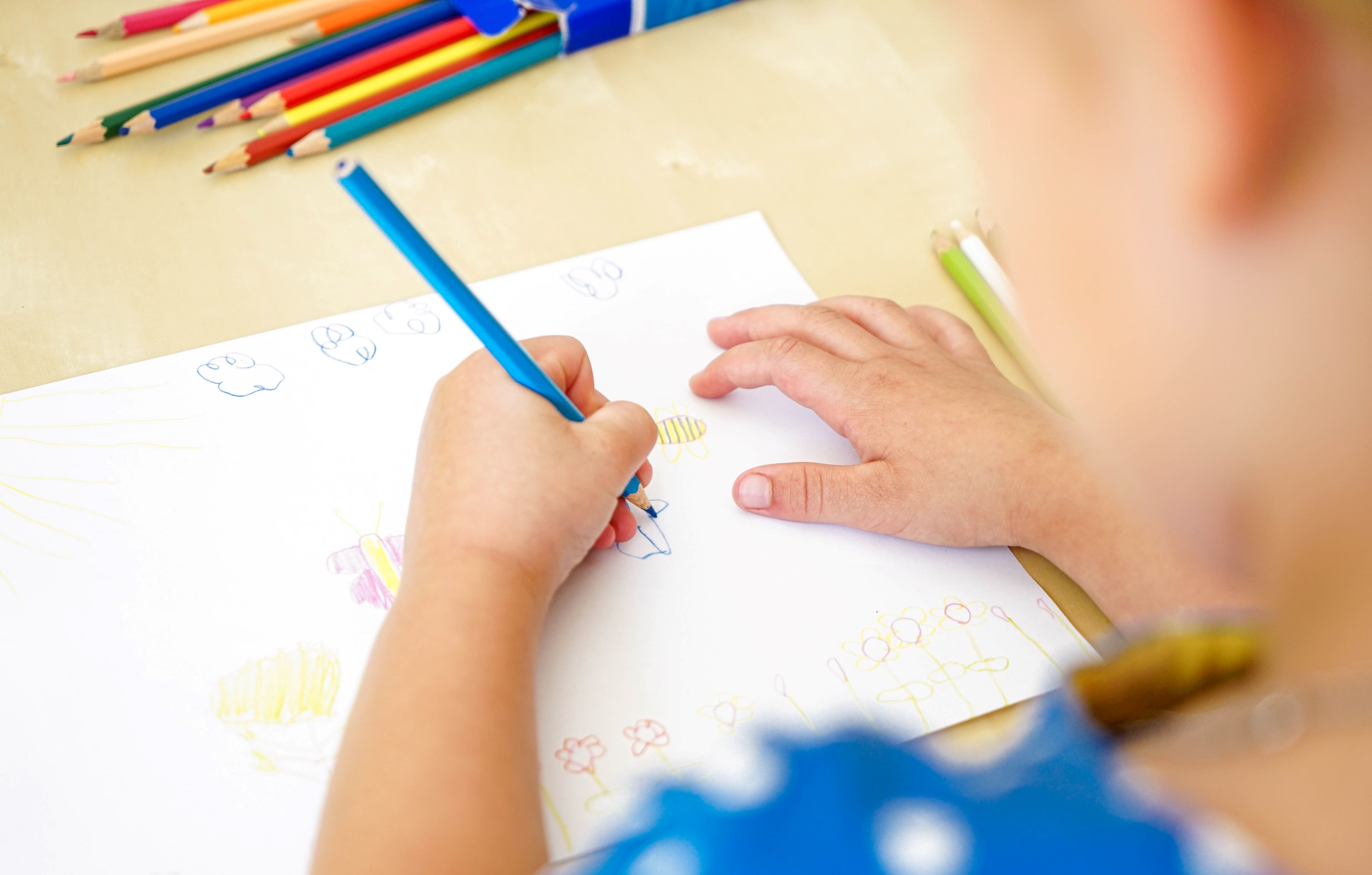 Hier malt ein kleines Kind etwas mit einem blauen Stift ein weißes Papier.
