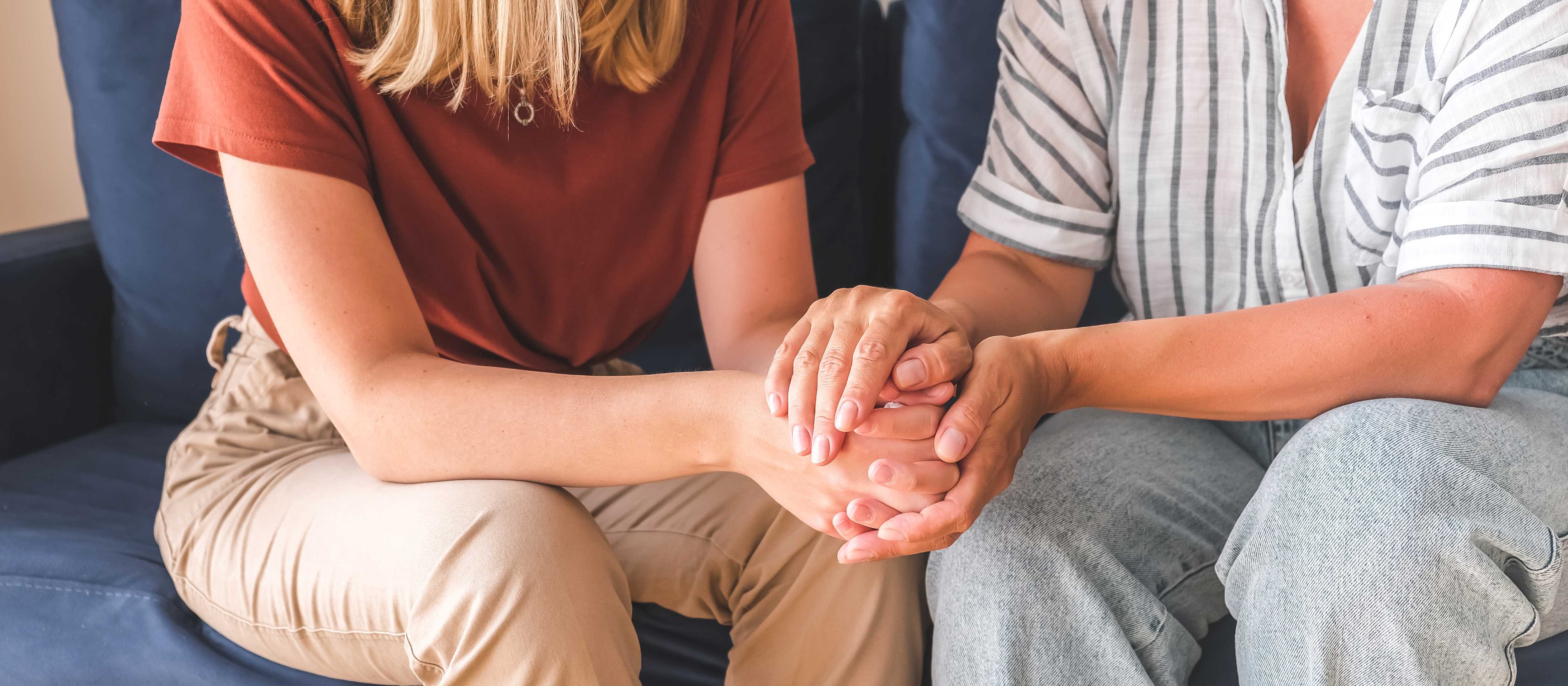 Zwei Menschen die nebeneinander sitzen und die Hände tröstend halten